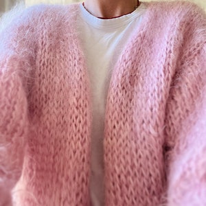 May include: A soft, pink fuzzy cardigan with a knitted pattern. The cardigan is open in the front and has long sleeves.