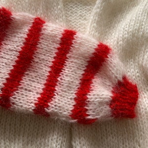 May include: White cardigan with red stripes on the sleeves. The cardigan is made of a soft, fuzzy yarn.