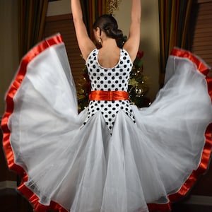 May include: A white and black polka dot dance dress with a flowing skirt and red trim. The dress has a red sash and is being worn with arms raised. The dress is suitable for ballroom or other dance performances.