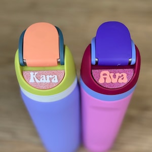 May include: Two colorful water bottles with personalized names, "Kara" and "Ava", in pink and white letters on a glittery background. The bottles are purple and pink with a light blue lid.