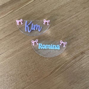May include: Two clear, semi-circular name tags with pink bows. One tag has the name "Kim" in lavender script, and the other has "Romina" in light blue script. The tags are on a wooden surface.