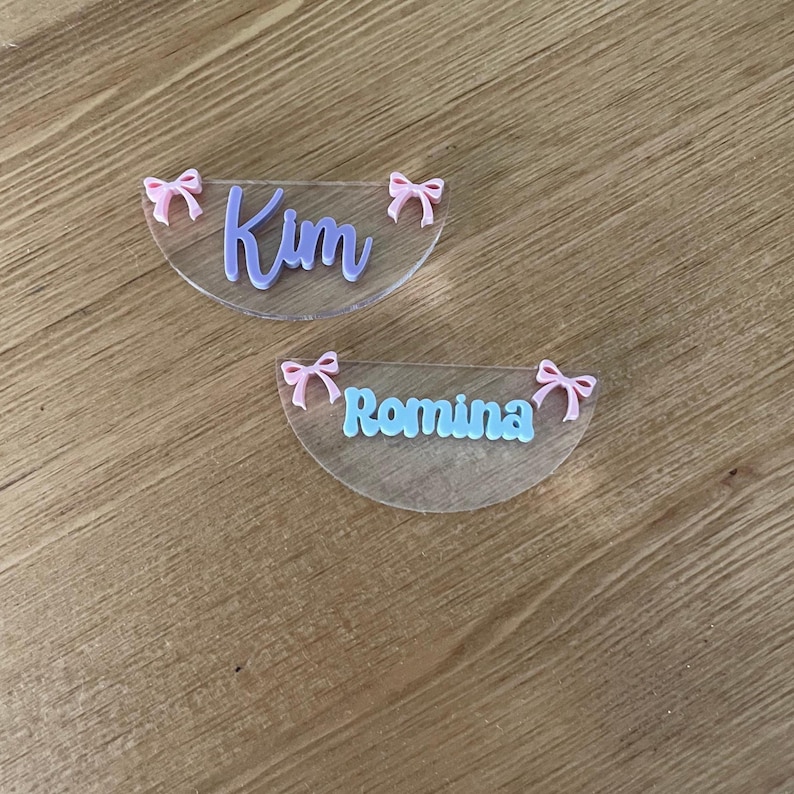 May include: Two clear, semi-circular name tags with pink bows. One tag has the name "Kim" in lavender script, and the other has "Romina" in light blue script. The tags are on a wooden surface.