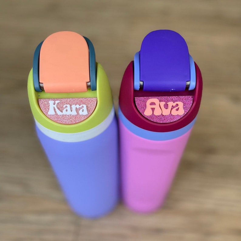 May include: Two colorful water bottles with personalized names, "Kara" and "Ava", in pink and white letters on a glittery background. The bottles are purple and pink with a light blue lid.