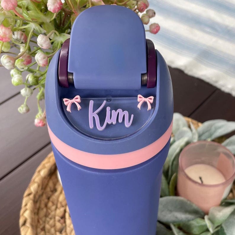 May include: A blue water bottle with a pink accent band and a flip-top lid. The name "Kim" is displayed in a light purple script with pink bow accents. The bottle is set against a blurred background.