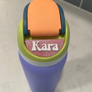 May include: A purple water bottle with a green, white, and orange lid. The lid has a flip top and a pink glitter sticker with the name "Kara" on it.