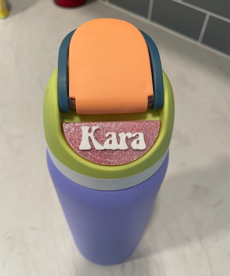May include: A purple water bottle with a green, white, and orange lid. The lid has a flip top and a pink glitter sticker with the name "Kara" on it.