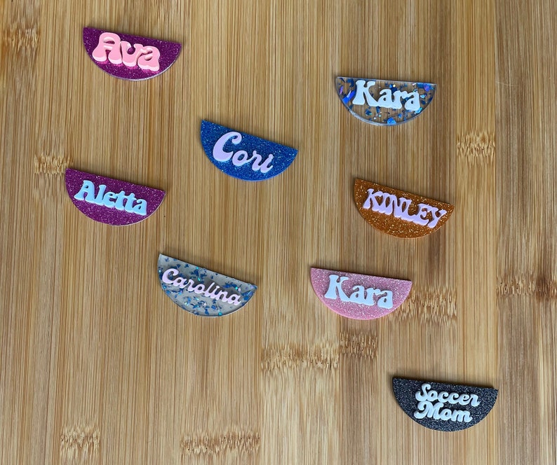 May include: Nine half-circle resin ornaments with glitter and names written in white. The names are Ava, Aletta, Cori, Kara, Kinley, Carolina, Kara, Soccer Mom.