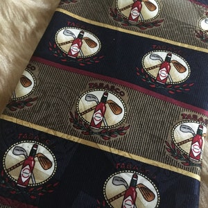 May include: A dark blue silk tie with a repeating pattern of gold and brown stripes. The pattern features a red Tabasco sauce bottle with two crossed golf clubs in a circle. The text "TABASCO" is printed in gold letters above the bottle.