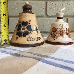 May include: Two ceramic bells with floral designs. The bell on the left is brown with a blue flower and says "Cozumel". The bell on the right is white with brown flowers and says "Cancun".