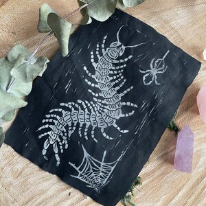 Pode incluir: Uma peça de arte têxtil preta com um design branco de centopeia, aranha e teia de aranha. A obra de arte é exibida em uma superfície de madeira com folhas de eucalipto e um cristal rosa. A centopeia e a aranha são o ponto focal do design.