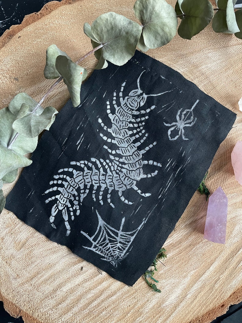 May include: A black textile art piece with a white centipede, spider, and spiderweb design. The artwork is displayed on a wooden surface with eucalyptus leaves and a pink crystal. The centipede and spider are the focal point of the design.