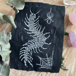 May include: A black fabric art piece with a white linocut print of a centipede, spider, and spiderweb. The artwork has a handmade, rustic feel. It is displayed on a wooden surface, with eucalyptus leaves and crystals adding to the aesthetic.