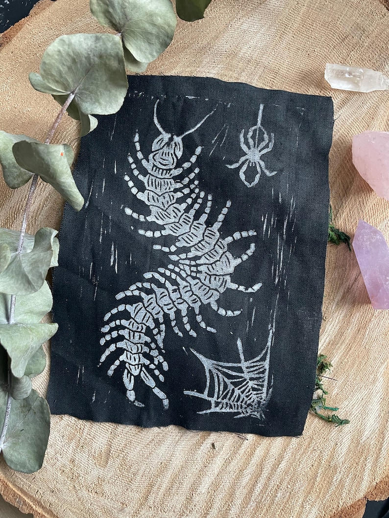 May include: A black fabric art piece with a white linocut print of a centipede, spider, and spiderweb. The artwork has a handmade, rustic feel. It is displayed on a wooden surface, with eucalyptus leaves and crystals adding to the aesthetic.