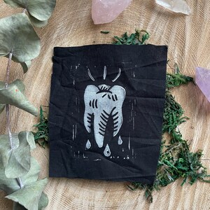 以下が含まれることがあります： 白いインクで歯が描かれた黒い布のパッチ。歯から3滴の液体が落ちている。歯は線と点でスタイリッシュにデザインされている
