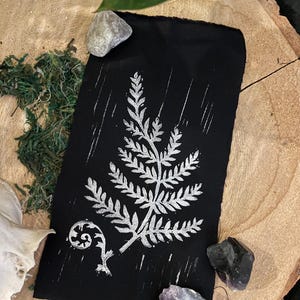 May include: A black fabric print featuring a silver fern design. The print is on a black background and includes a fern with a curled frond.