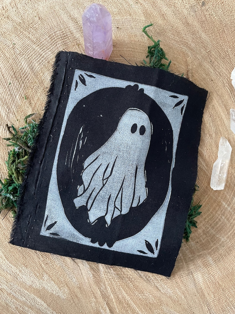 以下が含まれることがあります： 白い円の中に白い幽霊のイラストが描かれた黒い布のパッチ。幽霊は2つの目とシンプルで流れるような形をしています。