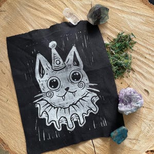 Peut inclure: Un tissu carré noir avec une impression linogravure blanche d'un chat portant un chapeau de fête et un col à volants. Le chat a de grands yeux et un petit sourire. Le tissu est sur une surface en bois avec des cristaux et de la mousse.