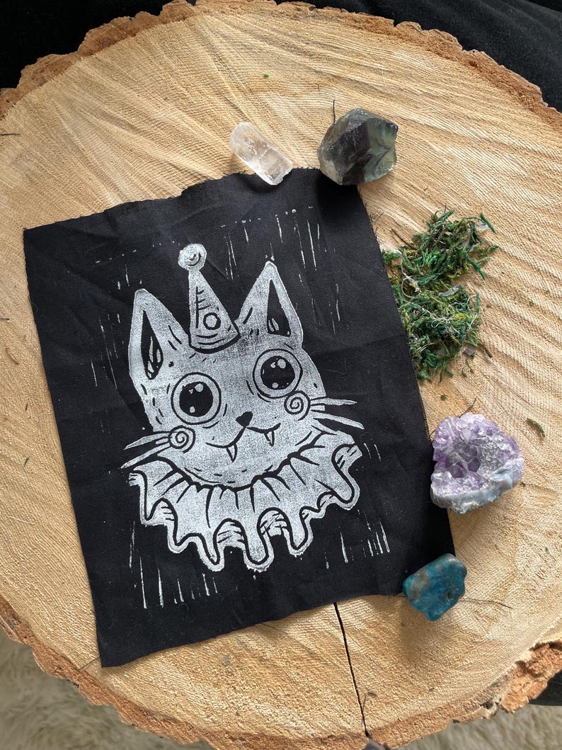 May include: A black square fabric with a white linocut print of a cat wearing a party hat and ruffled collar. The cat has large eyes and a small smile. The fabric is on a wooden surface with crystals and moss.