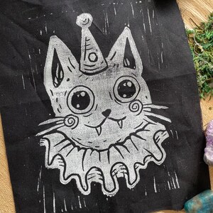May include: Black fabric patch featuring a whimsical cat design. The cat has large eyes, a party hat, and a ruffled collar, all in white. The design has a hand-carved, linocut style. The patch is likely for sewing or ironing onto clothing or accessories.