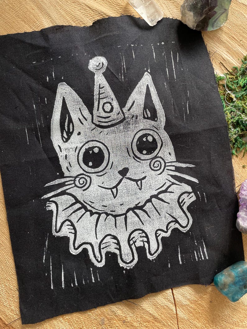 May include: Black fabric patch featuring a whimsical cat design. The cat has large eyes, a party hat, and a ruffled collar, all in white. The design has a hand-carved, linocut style. The patch is likely for sewing or ironing onto clothing or accessories.