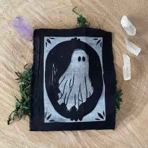 以下が含まれることがあります： 白い楕円形の枠の中に白い幽霊のイラストが描かれた黒い布のパッチ。幽霊は2つの目とシンプルで流れるような形をしています。枠には装飾的な縁取りがあります。