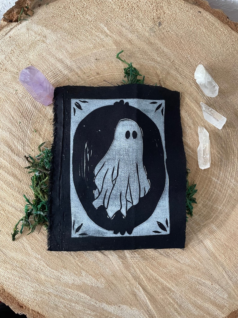 以下が含まれることがあります： 白い楕円形の枠の中に白い幽霊のイラストが描かれた黒い布のパッチ。幽霊は2つの目とシンプルで流れるような形をしています。枠には装飾的な縁取りがあります。