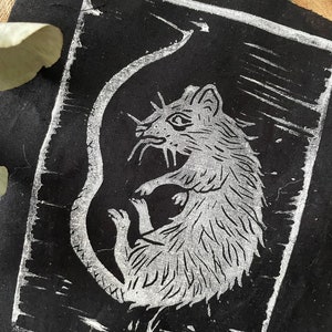 Peut inclure: Un patch en tissu noir avec un contour blanc, repr&eacute;sentant un rat en ligne blanche avec une longue queue enroul&eacute;e autour de son corps.