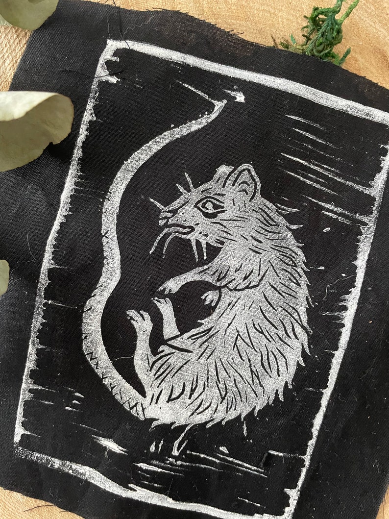 Peut inclure: Un patch en tissu noir avec un contour blanc, repr&eacute;sentant un rat en ligne blanche avec une longue queue enroul&eacute;e autour de son corps.