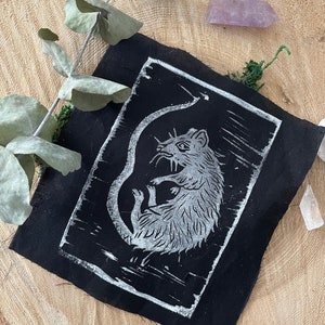 Peut inclure: Un patch en tissu noir avec une impression linogravure blanche d'un rat. Le rat est enroul&eacute; sur lui-m&ecirc;me, sa queue enroul&eacute;e autour de son corps. L'impression est sur un fond noir.