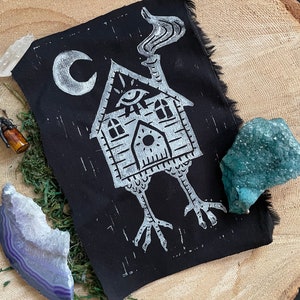 May include: Black fabric patch with a white print of a house with a single eye, a chimney with smoke, and two legs. The house has a crescent moon above it.