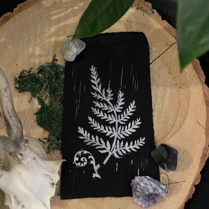 May include: A black fabric print with a white fern design. The print is on a wooden surface with a small piece of moss and a few crystals.