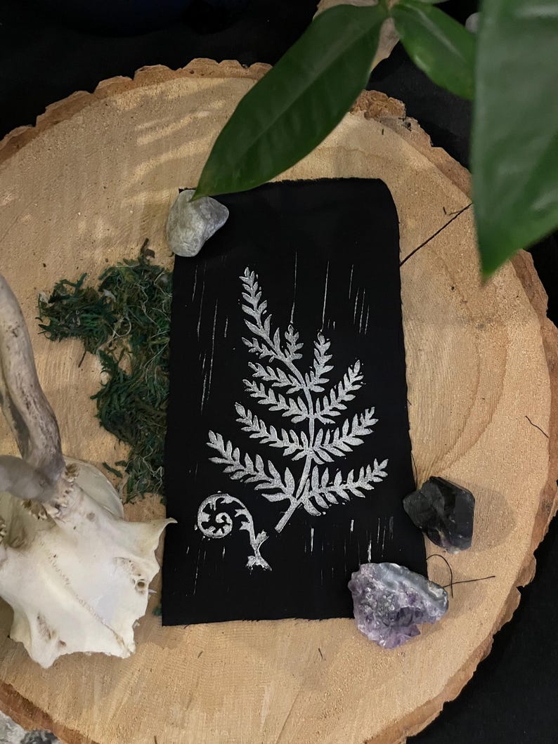 May include: A black fabric print with a white fern design. The print is on a wooden surface with a small piece of moss and a few crystals.