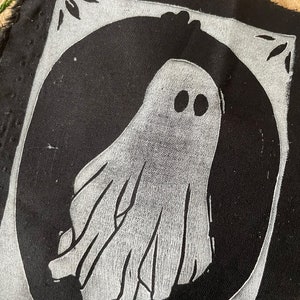 以下が含まれることがあります： 白い幽霊のデザインが施された黒い布のパッチ。幽霊は2つの目と流れるような形をしています。パッチは白い縁取りで囲まれています。
