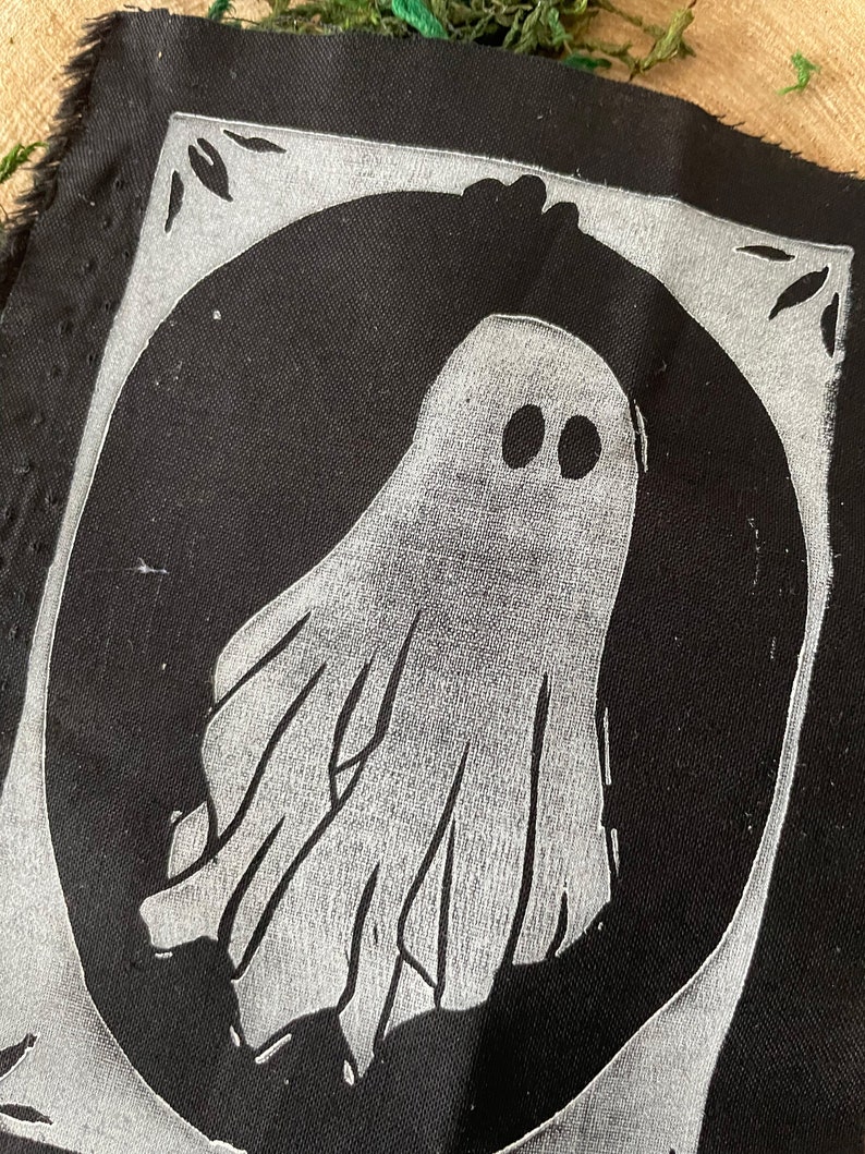 以下が含まれることがあります： 白い幽霊のデザインが施された黒い布のパッチ。幽霊は2つの目と流れるような形をしています。パッチは白い縁取りで囲まれています。