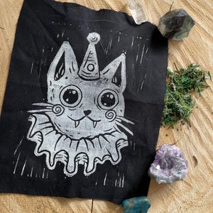 May include: A black fabric patch featuring a white, whimsical cat design. The cat wears a party hat and a ruffled collar. The patch is surrounded by crystals and moss on a wooden surface.