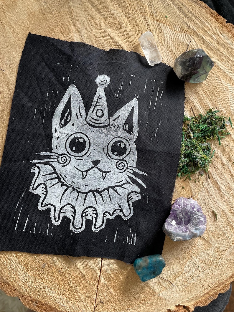 May include: A black fabric patch featuring a white, whimsical cat design. The cat wears a party hat and a ruffled collar. The patch is surrounded by crystals and moss on a wooden surface.