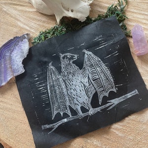 Könnte beinhalten: Ein schwarzer Stoff-Patch mit einem weißen Aufdruck einer Fledermaus mit ausgebreiteten Flügeln. Die Fledermaus sitzt auf einem Ast.
