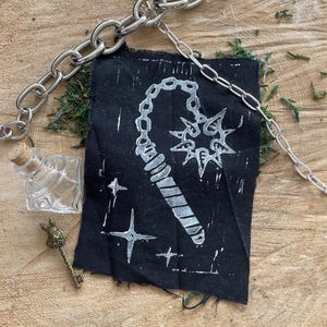 Op de afbeelding: Een zilveren ketting en een zwart stoffen lapje met een witte illustratie van een dorsvlegel en sterren. Een kleine glazen fles en een messing sleutel zijn ook aanwezig, alles gerangschikt op een houten oppervlak met mos.