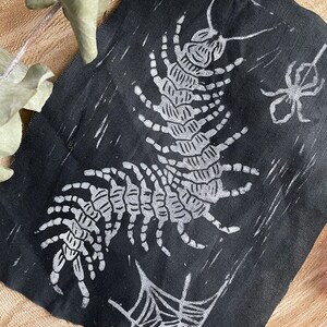 May include: Black fabric featuring a white centipede design, a spider, and a spiderweb. The centipede is the main element, with a detailed segmented body and legs. The spider and web are smaller, supporting designs. The fabric has a slightly textured appearance.