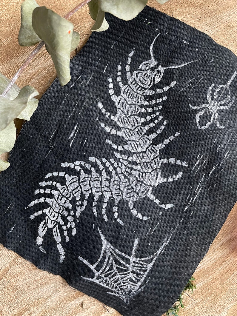 May include: Black fabric featuring a white centipede design, a spider, and a spiderweb. The centipede is the main element, with a detailed segmented body and legs. The spider and web are smaller, supporting designs. The fabric has a slightly textured appearance.