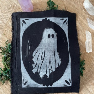以下が含まれることがあります： 白い幽霊のデザインが円の中に描かれた黒い布のパッチ。幽霊は2つの目とシンプルで流れるような形をしています。パッチは白い縁取りと装飾的な要素で囲まれています。