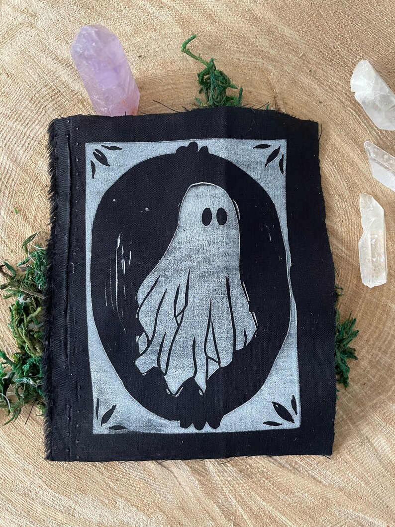 以下が含まれることがあります： 白い幽霊のデザインが円の中に描かれた黒い布のパッチ。幽霊は2つの目とシンプルで流れるような形をしています。パッチは白い縁取りと装飾的な要素で囲まれています。