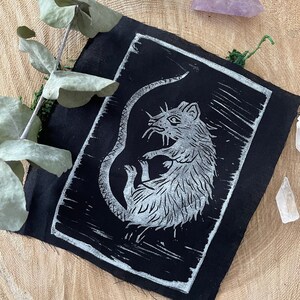 Peut inclure: Un patch en tissu noir avec une impression linogravure blanche d'un rat. Le rat est enroul&eacute; sur lui-m&ecirc;me, sa queue enroul&eacute;e autour de son corps.