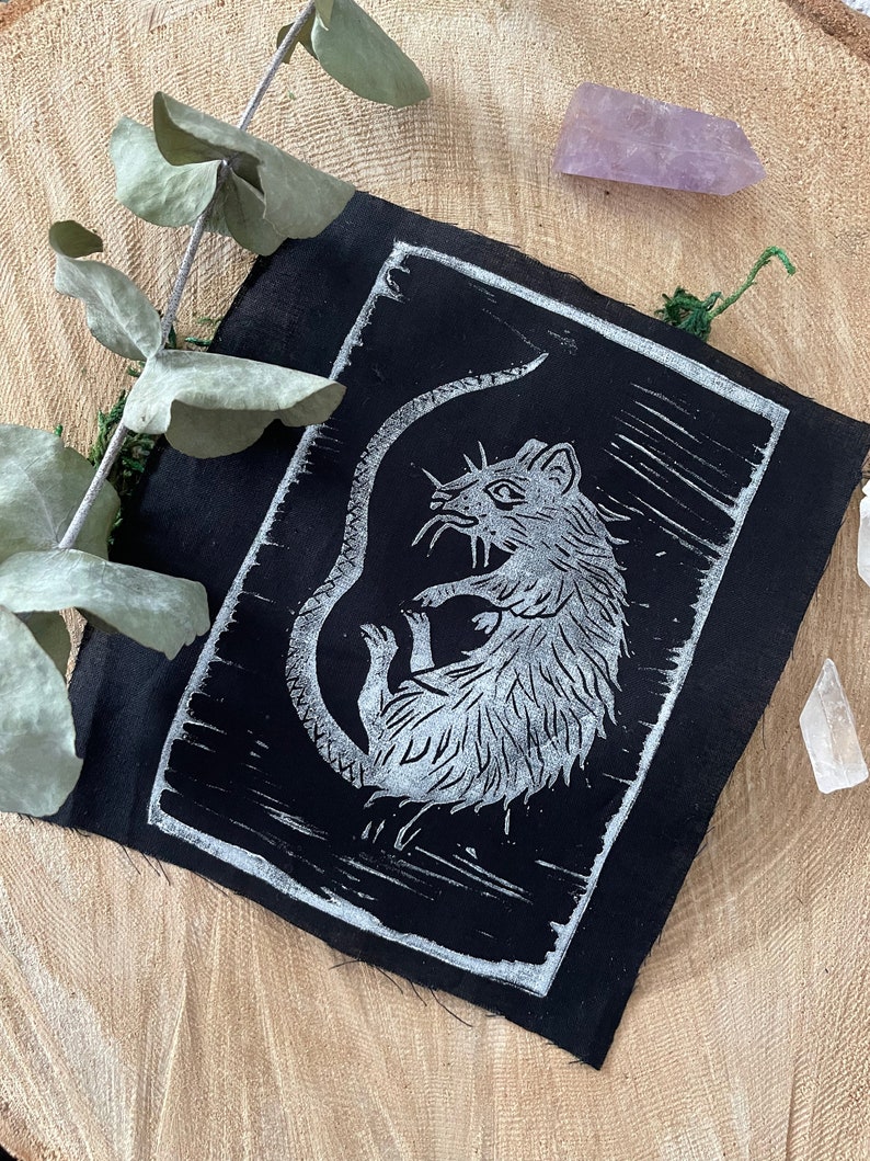 Peut inclure: Un patch en tissu noir avec une impression linogravure blanche d'un rat. Le rat est enroul&eacute; sur lui-m&ecirc;me, sa queue enroul&eacute;e autour de son corps.