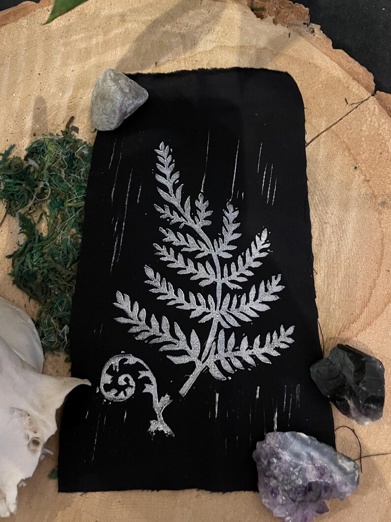 May include: A black fabric with a white fern design printed on it. The fabric is surrounded by small stones and moss.