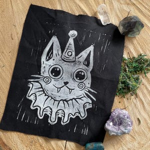 May include: A black fabric patch featuring a white illustration of a cat wearing a party hat and ruffled collar. The cat has large eyes and a smiling expression. The patch is surrounded by crystals and moss.