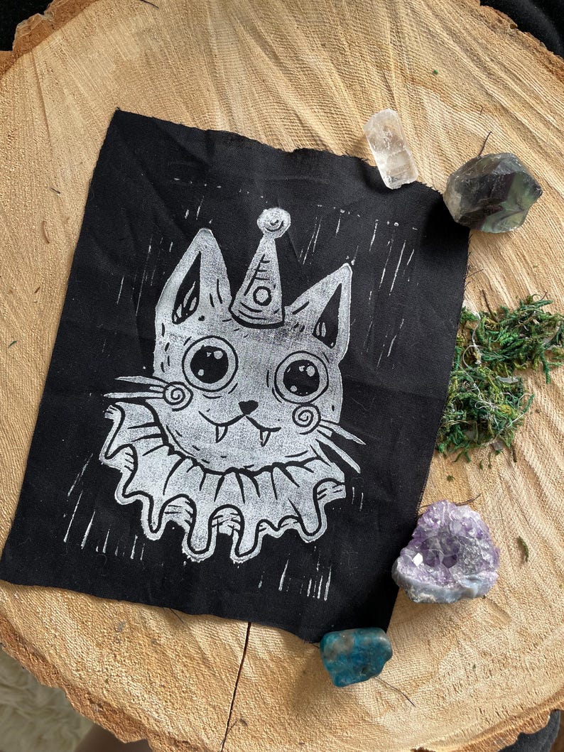 May include: A black fabric patch featuring a white illustration of a cat wearing a party hat and ruffled collar. The cat has large eyes and a smiling expression. The patch is surrounded by crystals and moss.