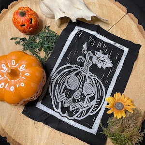 Può includere: Tessuto nero con una stampa a blocchi bianca di una zucca intagliata a forma di viso con uno stelo e una foglia. Il tessuto è incorniciato da un bordo bianco.