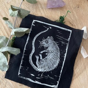 Peut inclure: Un patch en tissu noir avec une impression linogravure blanche d'un rat. Le rat est enroul&eacute; sur lui-m&ecirc;me, sa queue enroul&eacute;e autour de son corps. L'impression est sur un fond noir.