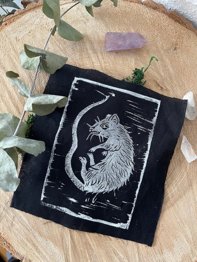 Peut inclure: Un patch en tissu noir avec une impression linogravure blanche d'un rat. Le rat est enroul&eacute; sur lui-m&ecirc;me, sa queue enroul&eacute;e autour de son corps. L'impression est sur un fond noir.